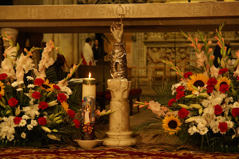 Colorful flower displays positioned on either side of a lit candle and a small statue of a golden arm with a finger pointing up.