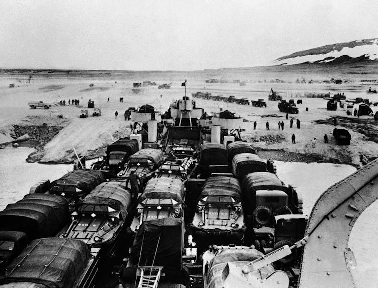 Trucks packed into a ship arrive with the ocean in the background.