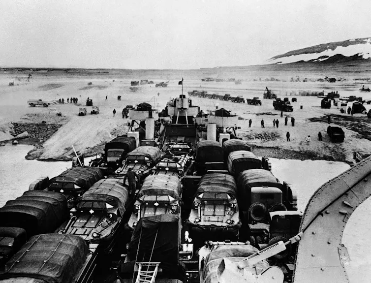 Trucks packed into a ship arrive with the ocean in the background.