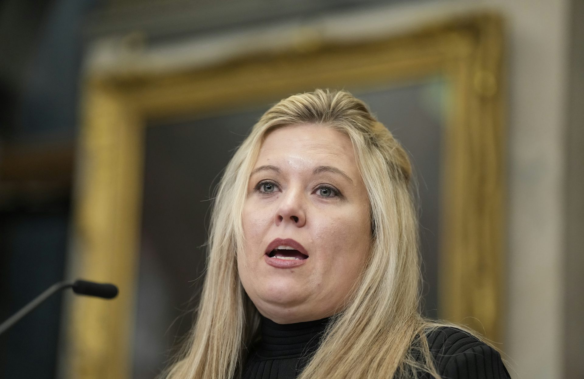A round-faced blonde woman speaks into a microphone.