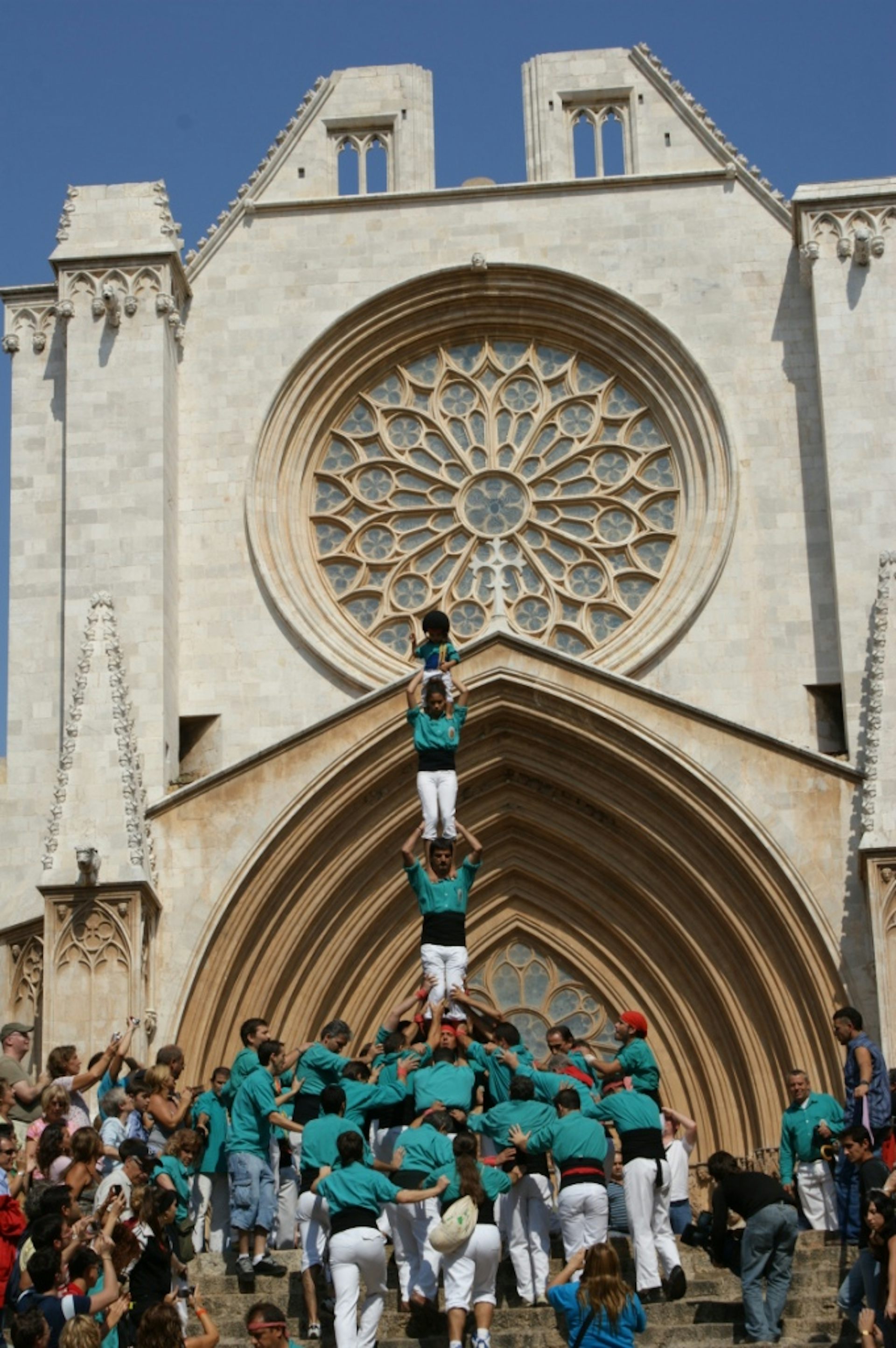People in white pants and teal shirts form a human pyramid in front of a cathedral with a large, circular stained-glass window.