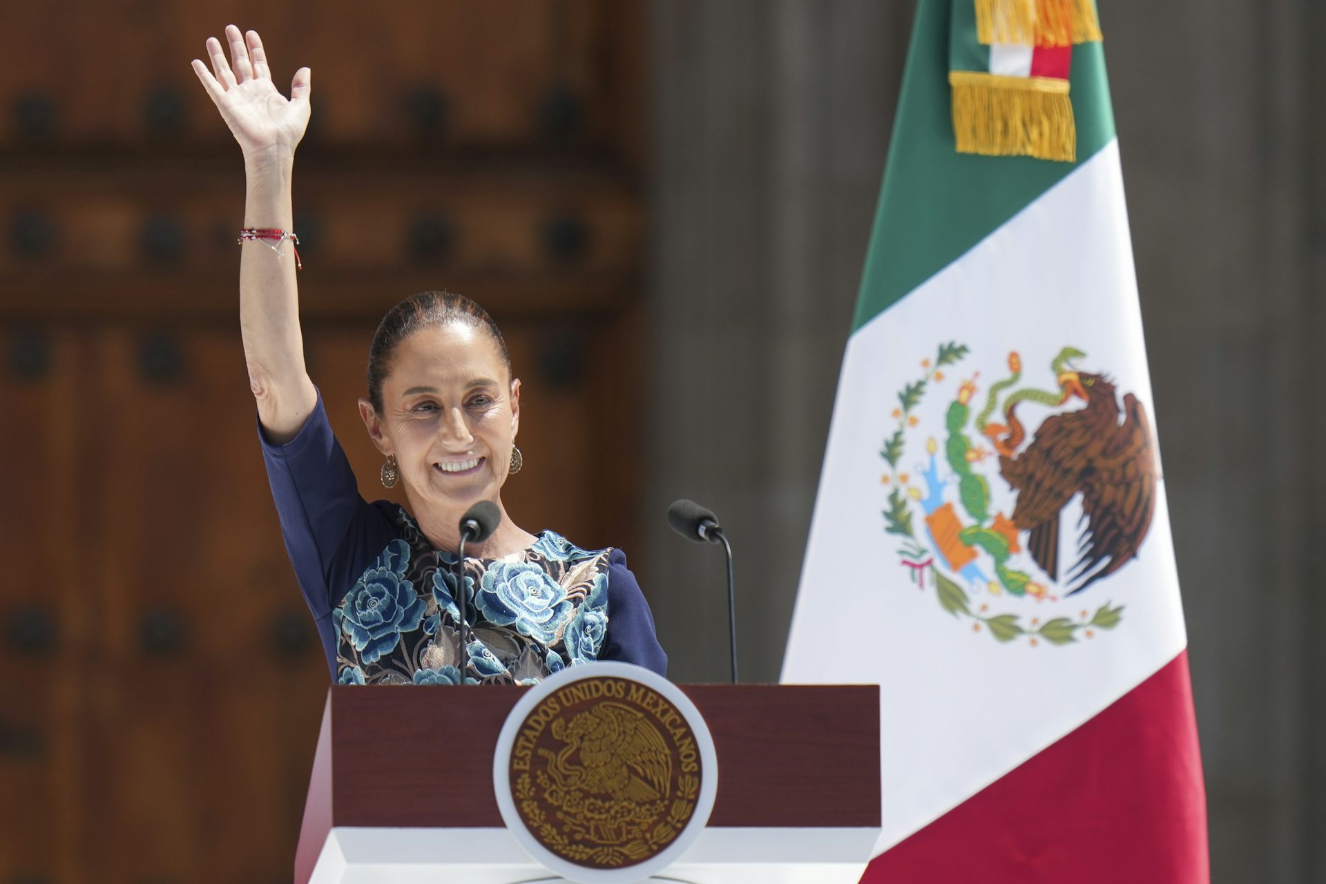 A woman waves from behind a podium with the Mexican flag standing beside her