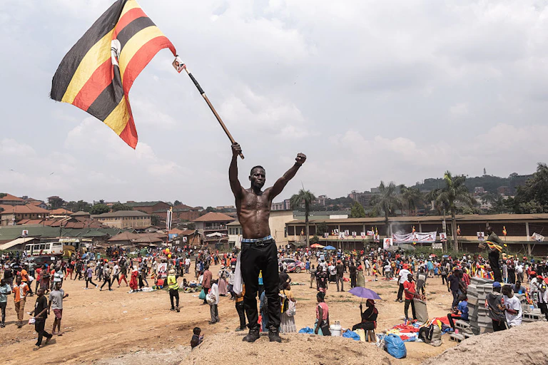 A man standing on a raised mound of soil holds a black, yellow and red flag while several people stand behind him