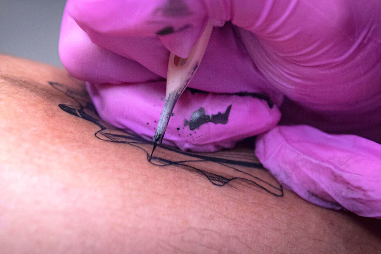 Close up of person in pink gloves making a black fine line tattoo with a single needle.