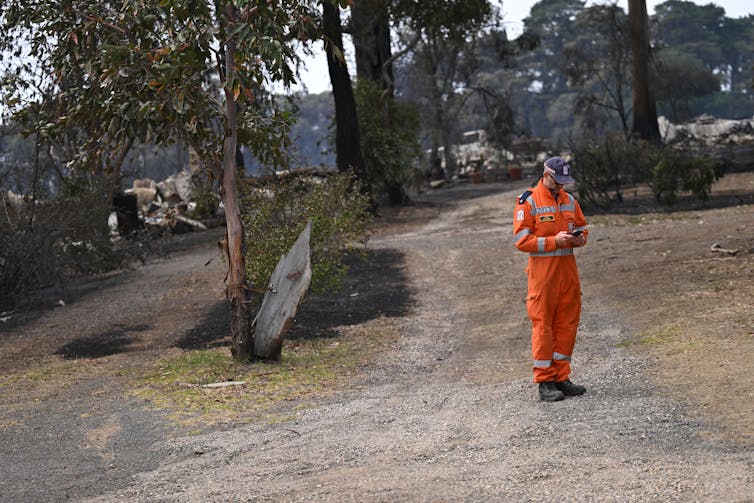 A member of the special emergency service, dressed in orange, looks at his phone in a burned landscape.