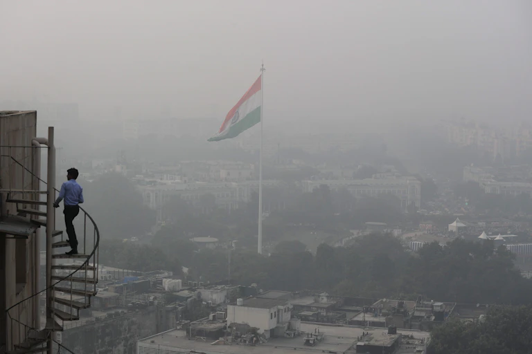 a large indian flag flies on a pole above a city enveloped in smog
