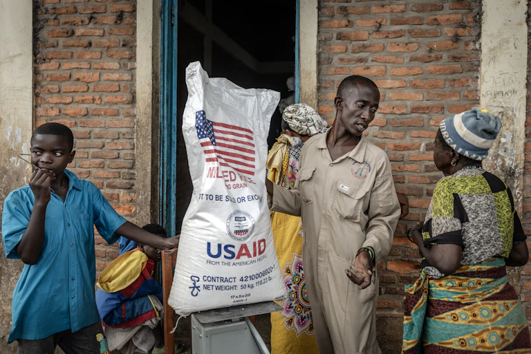 A man holds a sack of rice with USAID branding on it while speaking to other people.