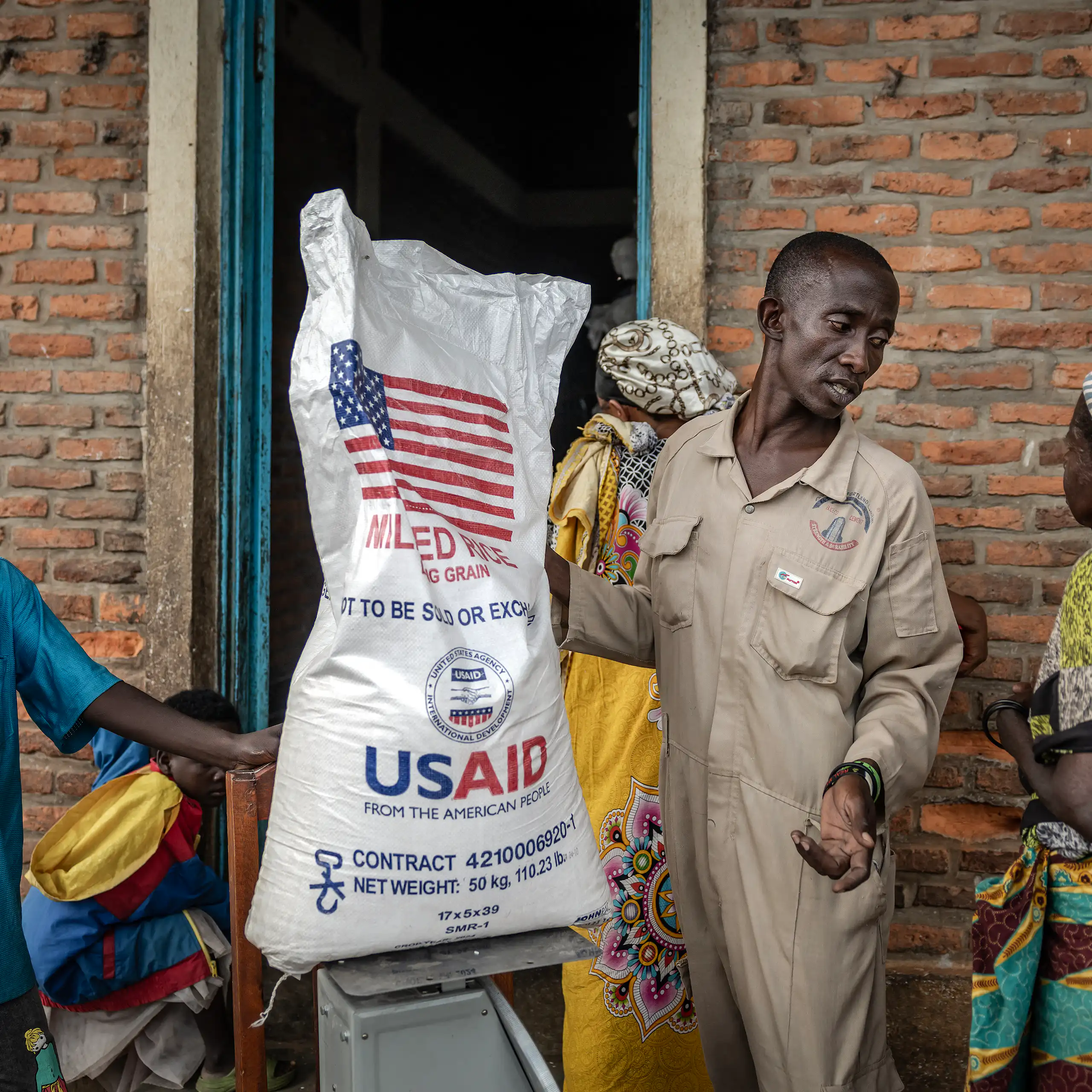 A man holds a sack of rice with USAID branding on it while speaking to other people.