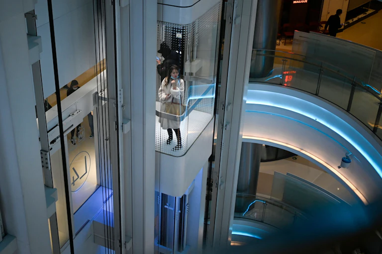 A young Asian woman peers out of a glass elevator in a shopping mall.