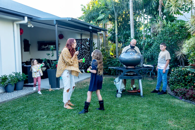 Mother blows bubbles with her daughters, whilst father and son cook on the barbeque