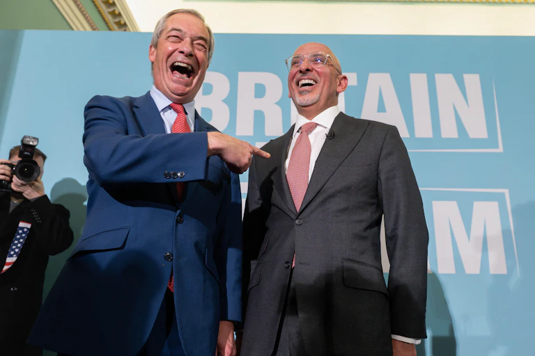 Nigel Farage stands beside Nadhim Zahawi and points to him, both men laughing, in front of a 'Britain needs reform' display