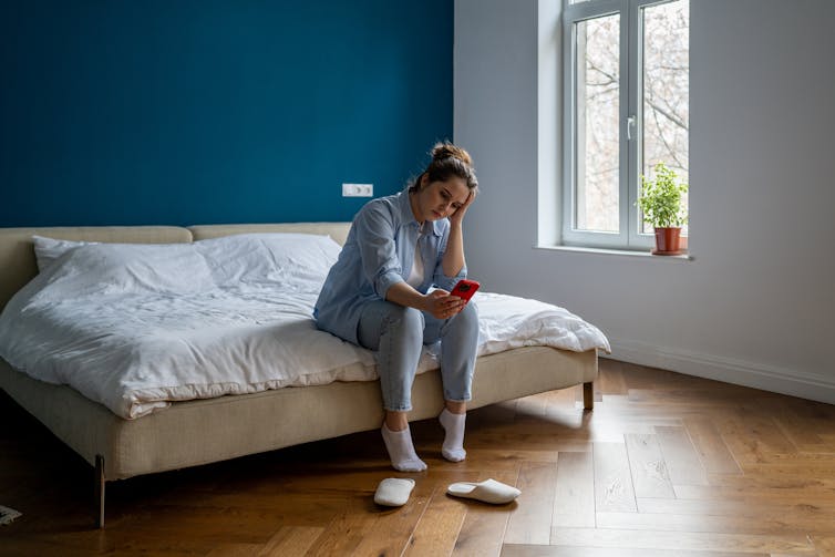 woman sits on end of bed holding head in hand while looking at phone