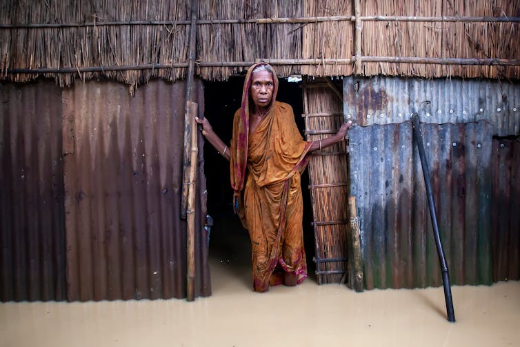 old woman stands at door of shack, flooded waters