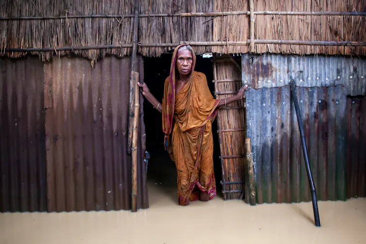 old woman stands at door of shack, flooded waters