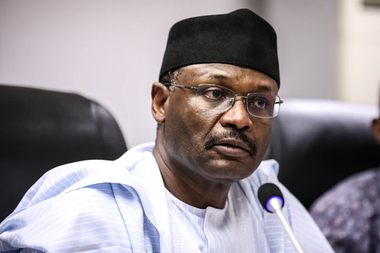 A man wearing a cap, African clothing and spectacles, with a microphone in front of him.