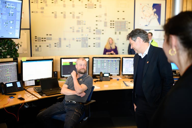 People are talking in a room with lots of computer screens and large diagrams on the wall.