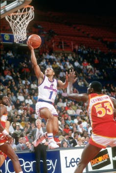 ¿Cómo conoce la gente sus intereses? El jugador más bajo de la NBA muestra cómo la confianza en uno mismo es más importante que la biología 1 Muggsy Bogues en el aire, con el brazo extendido hacia la red y una pelota de baloncesto en la mano, rodeado por jugadores del equipo competitivo.