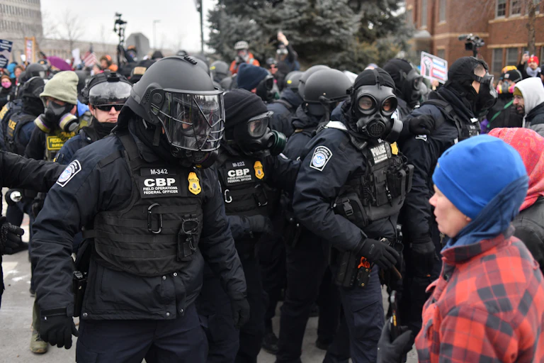 A group of law enforcement officers wearing helmets and masks, gathered outside a building with civilians facing them.