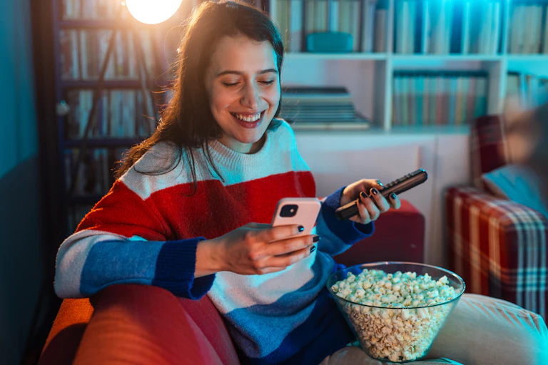 A young women on a couch with a bowl of popcorn, holding a TV remote and a smartphone and reading the phone screen.