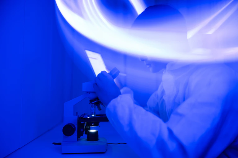 Scientist looking into microscope, their face blurred by a ribbon of light sweeping through a dark indigo room