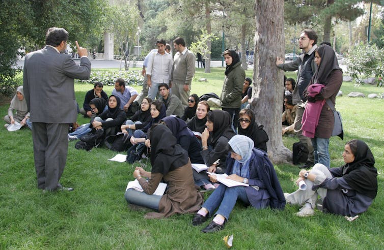 A group of young people, some of them with headscarves, sit on the grass near trees and look toward a man wearing a suit, with his finger in the air.