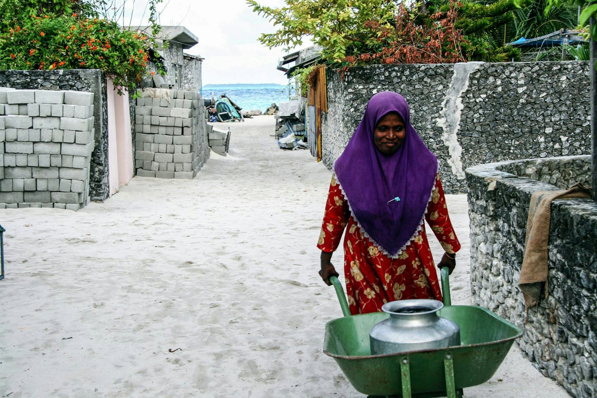 A woman pushes a wheelbarrow with a contain filled with freshwater. The ocean is behind her in the view.