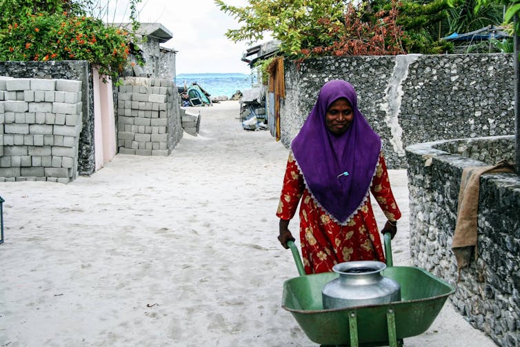 Una mujer empuja una carretilla con un recipiente lleno de agua dulce. El océano se ve detrás de ella.