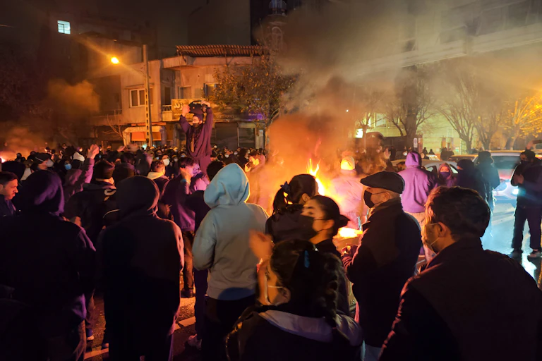 A large group of people are seen standing near a fire on the street at night.