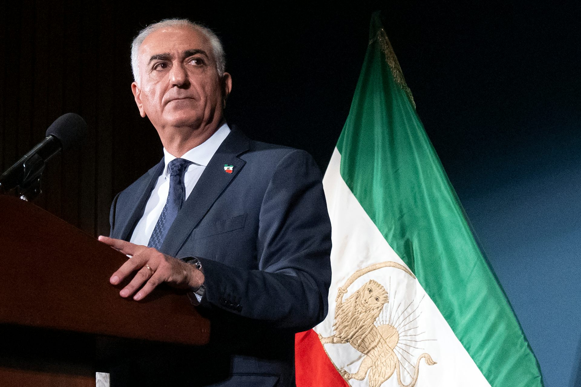Un hombre con cabello gris y traje se para frente a una bandera verde, blanca y roja.