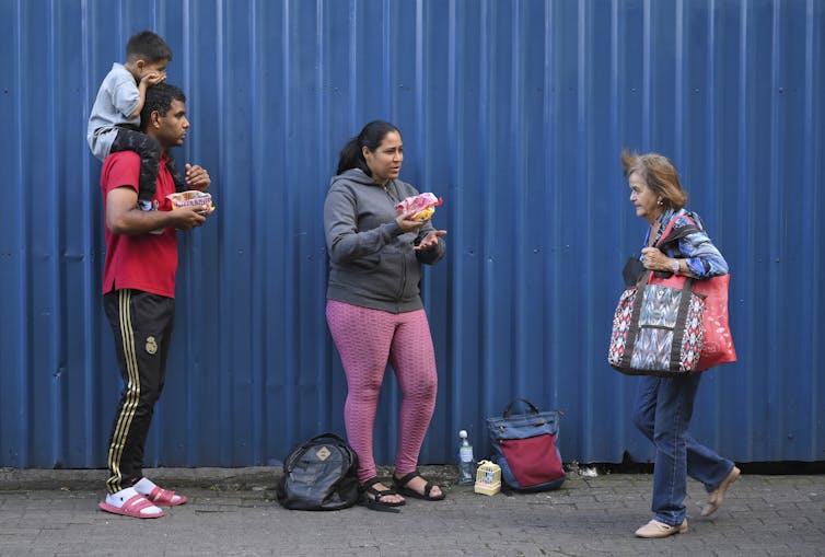 A man and woman stand on a street as a woman walks by. The man has a child on his shoulders.
