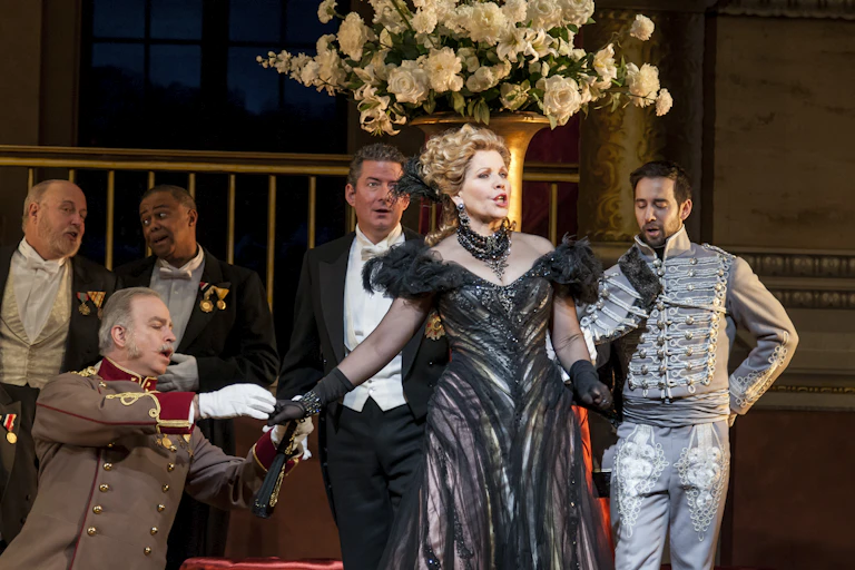 People dressed in dramatic costumes stand on stage, with a woman in a black dress singing.