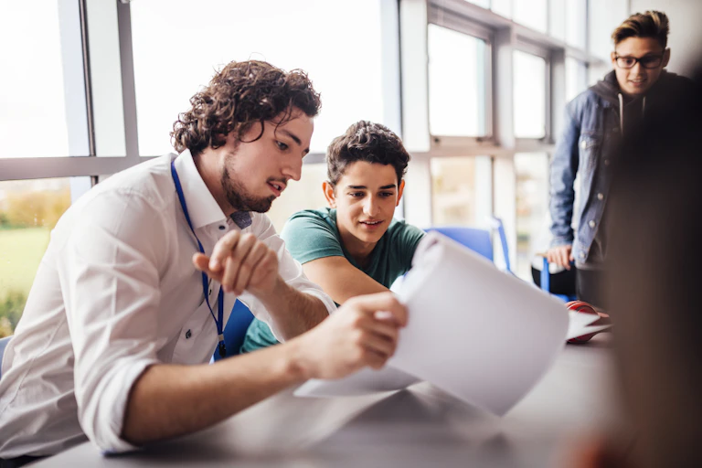 A teenager and young man sit at a desk. They are looking at sheets of paper and appear to be mid-conversation.