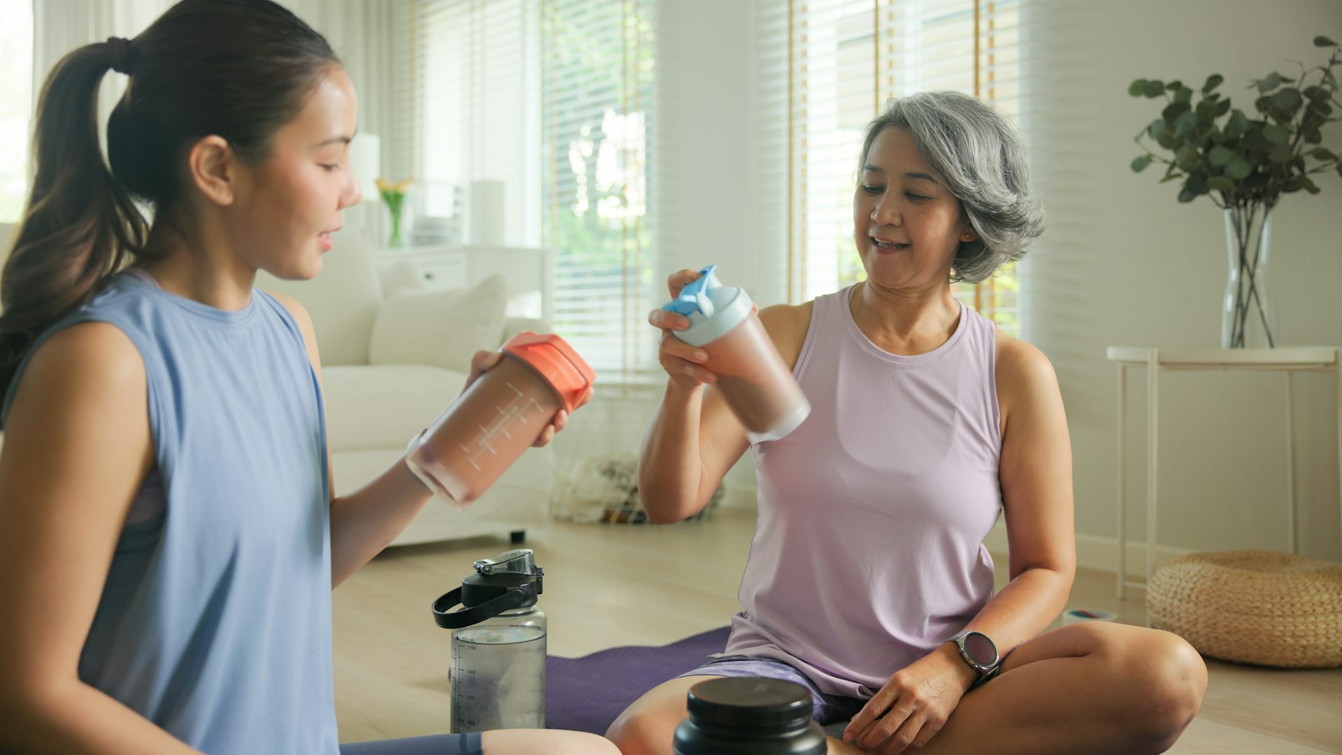 A young woman and an older woman sit on the floor on top of an exercise mat, each holding a shake bottle used for protein or creatine shakes.