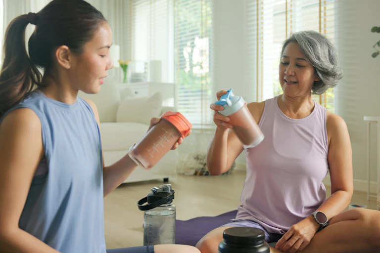 A young woman and an older woman sit on the floor on top of an exercise mat, each holding a shake bottle used for protein or creatine shakes.