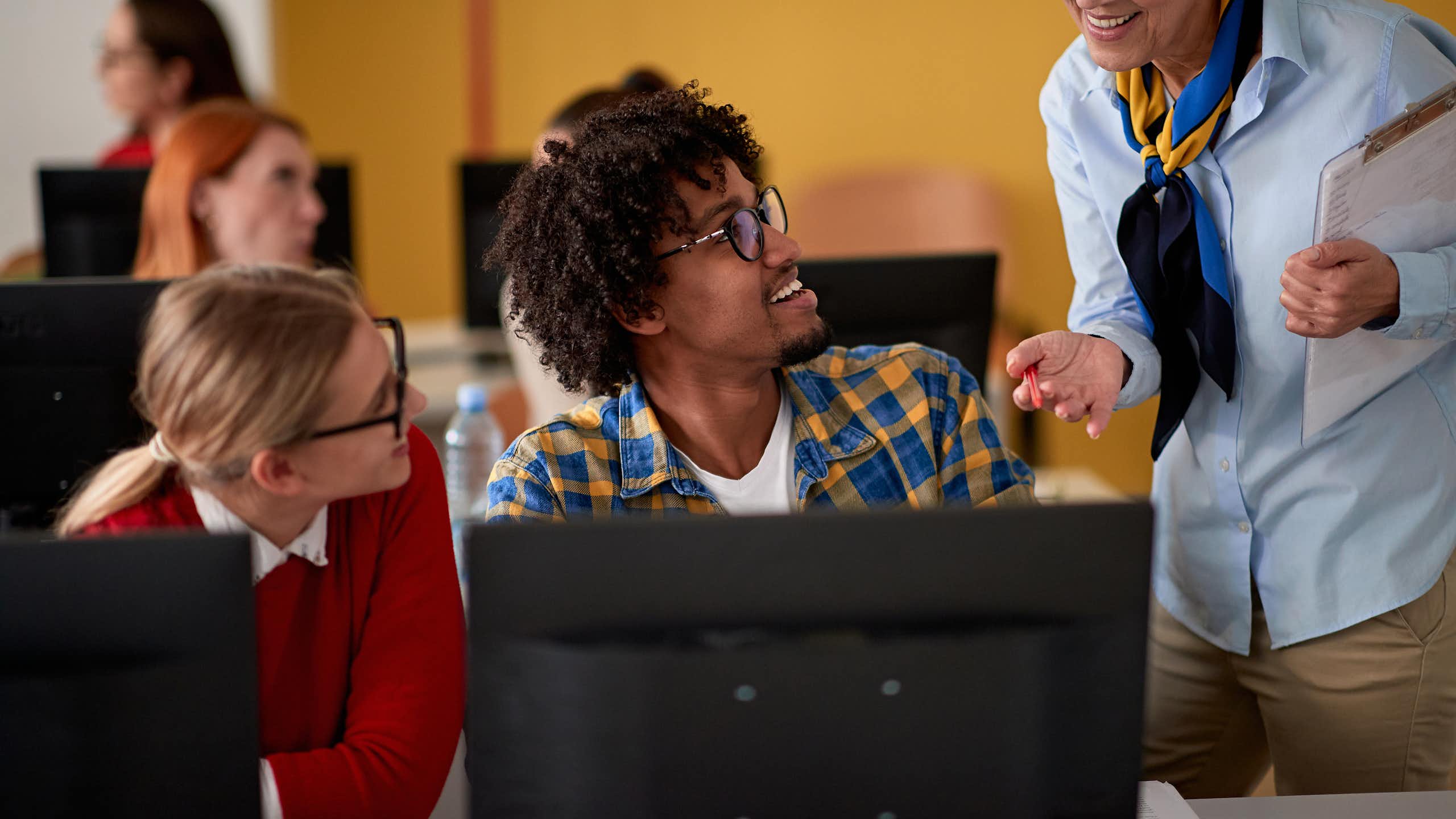 Una profesora sonriente se acerca para ver qué hacen dos jóvenes en sus ordenadores.