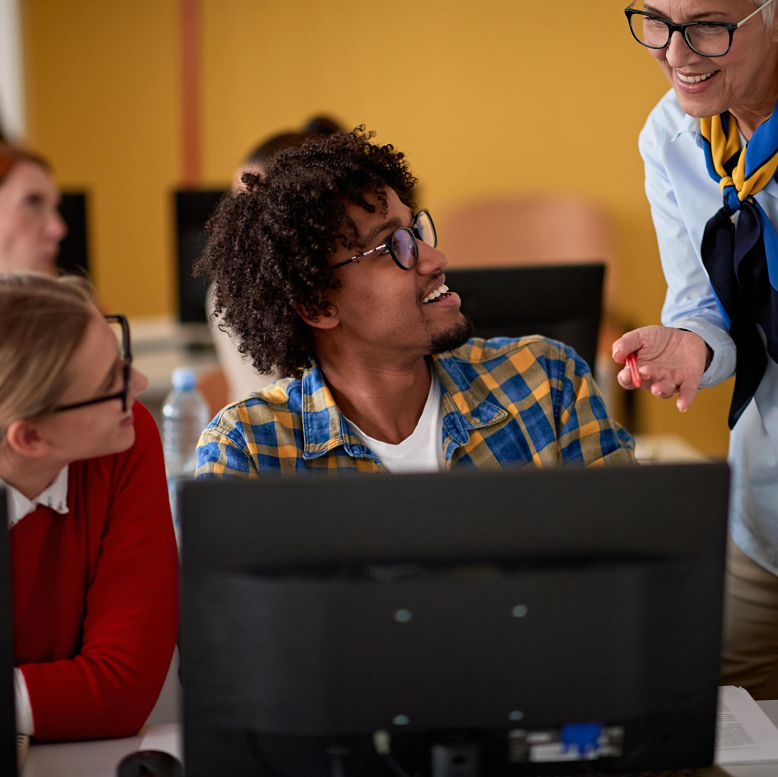 Una profesora sonriente se acerca para ver qué hacen dos jóvenes en sus ordenadores.