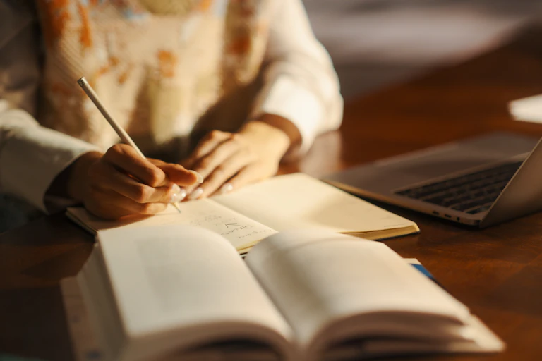 Hands writing notes from book and laptop