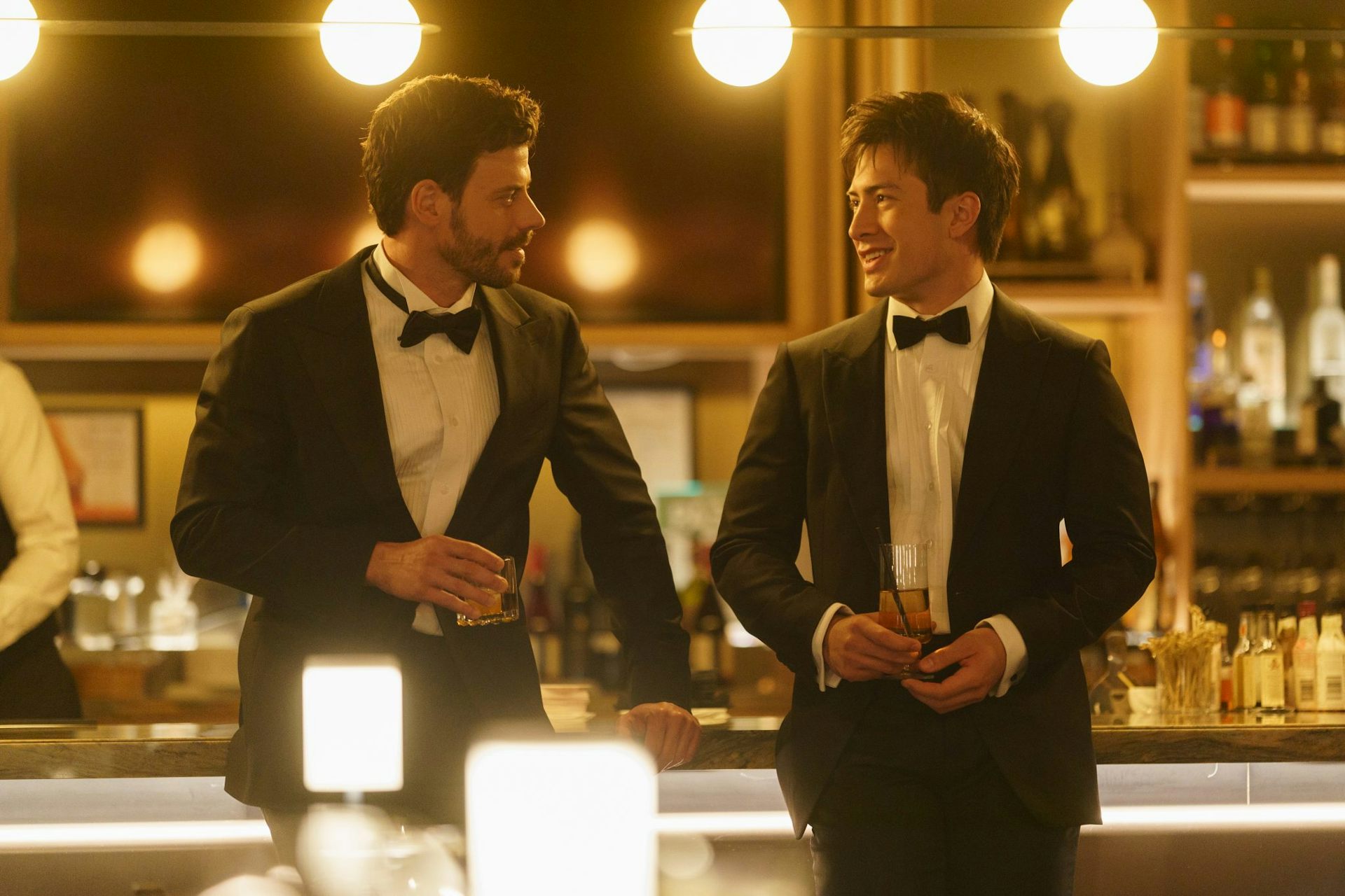 Two men in dressy suits leaning against a bar in a fancy environment.