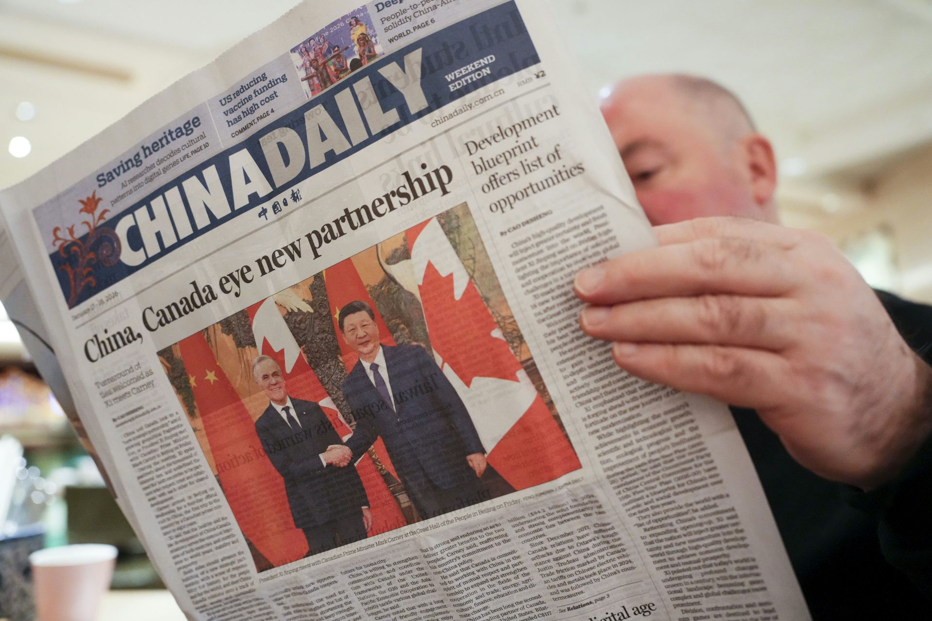 Un hombre leyendo un periódico con una foto de Mark Carney y Xi Jinping dándose la mano en el frente.
