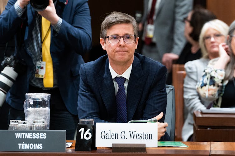 Un hombre blanco de mediana edad con gafas y traje se sienta detrás de una tarjeta con su nombre que dice Galen G. Weston.