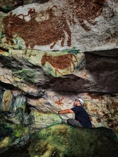 A vibrant image of a man in a white hard hat perched on rocks in a cave with large artworks above him.