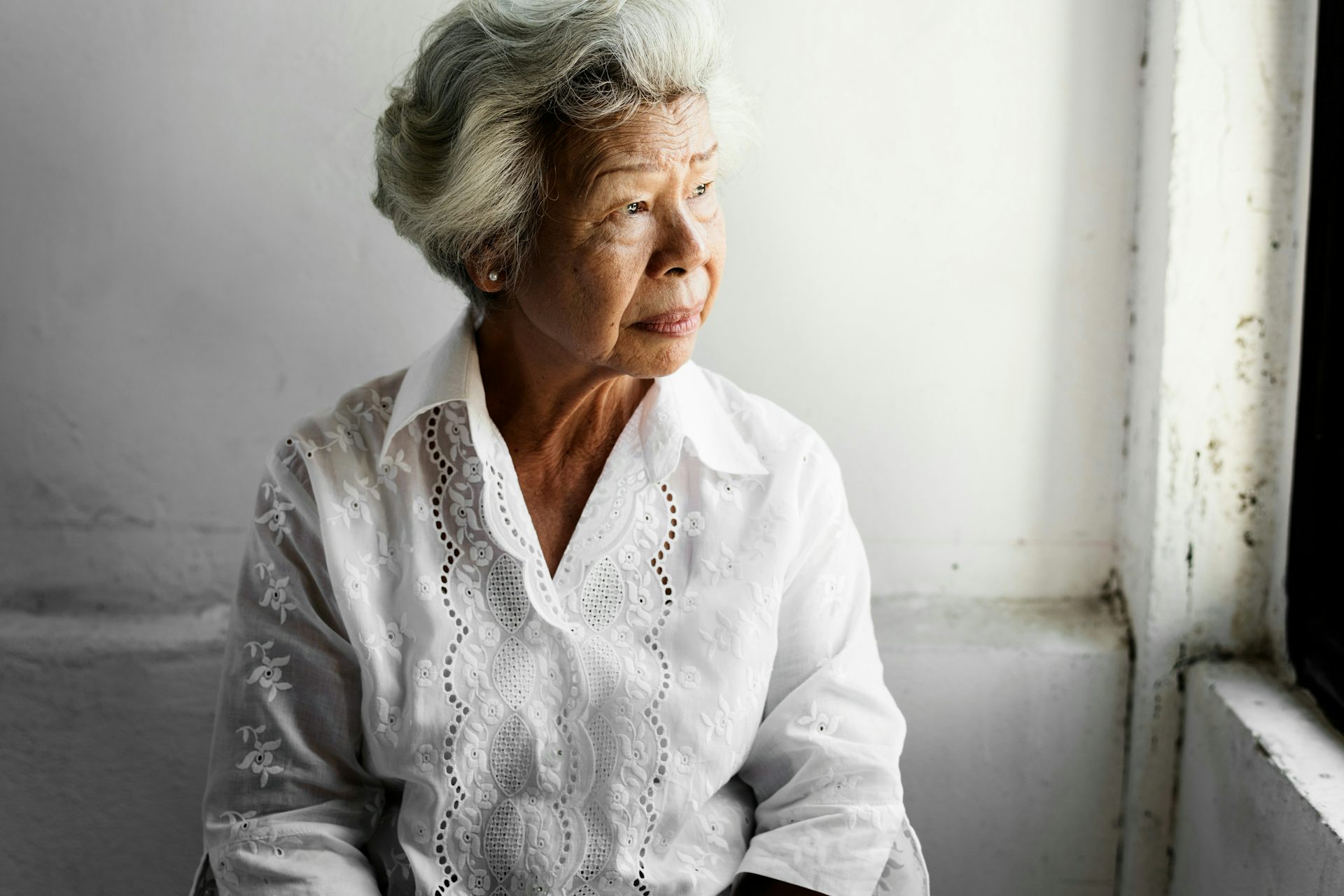 A woman with gray hair in a white blouse sitting at a window