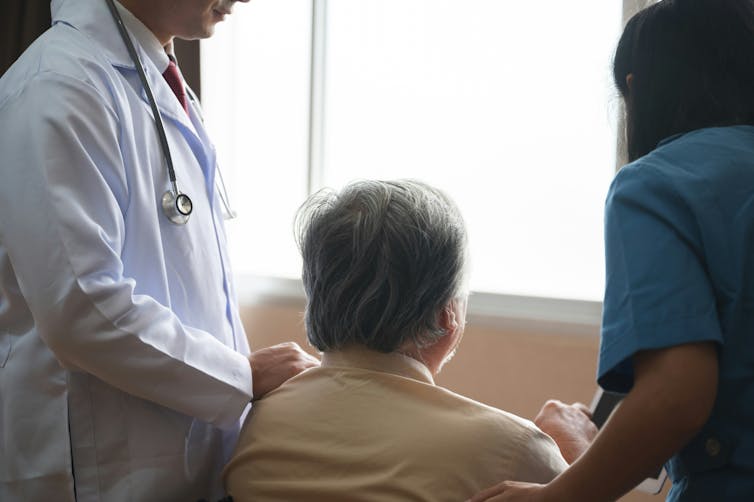 A man with gray hair seen from behind, seated with health-care workers on either side of him