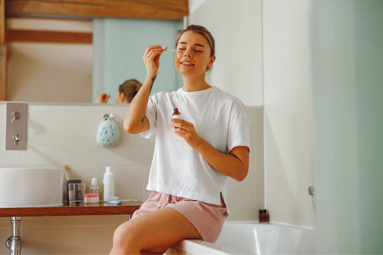 A young woman sitting on the edge of a bath tub applies a serum to her cheek.