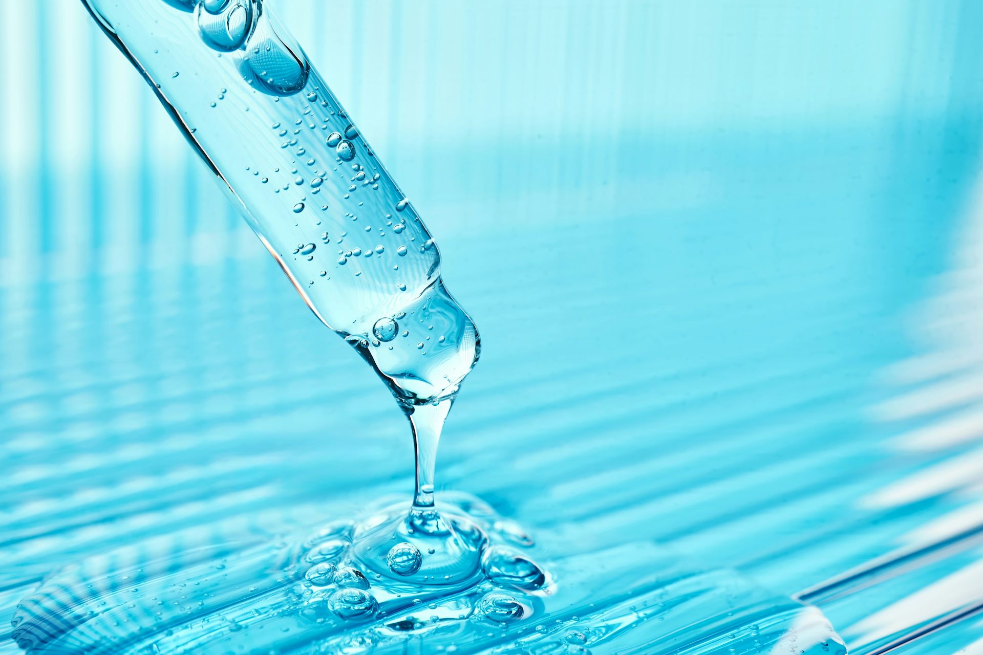 A dropper filled with blue liquid that is dripping out of the dropper and into a puddle of the same blue liquid.