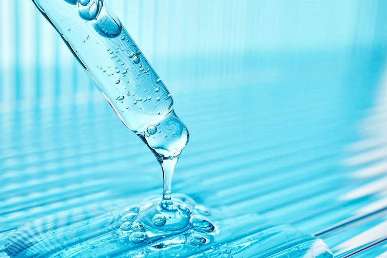 A dropper filled with blue liquid that is dripping out of the dropper and into a puddle of the same blue liquid.