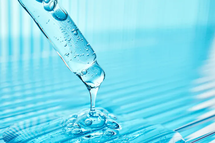 A dropper filled with blue liquid that is dripping out of the dropper and into a puddle of the same blue liquid.
