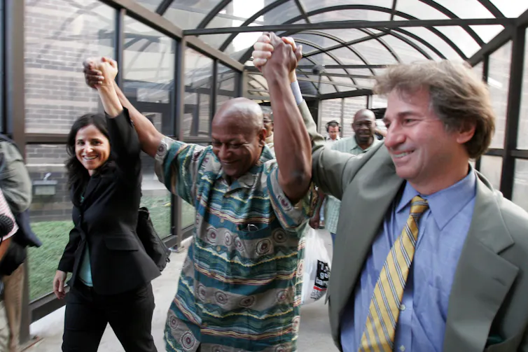 A Black man smiles and clasps his raised hands with a man with shaggy light brown hair and a dark-haired woman.