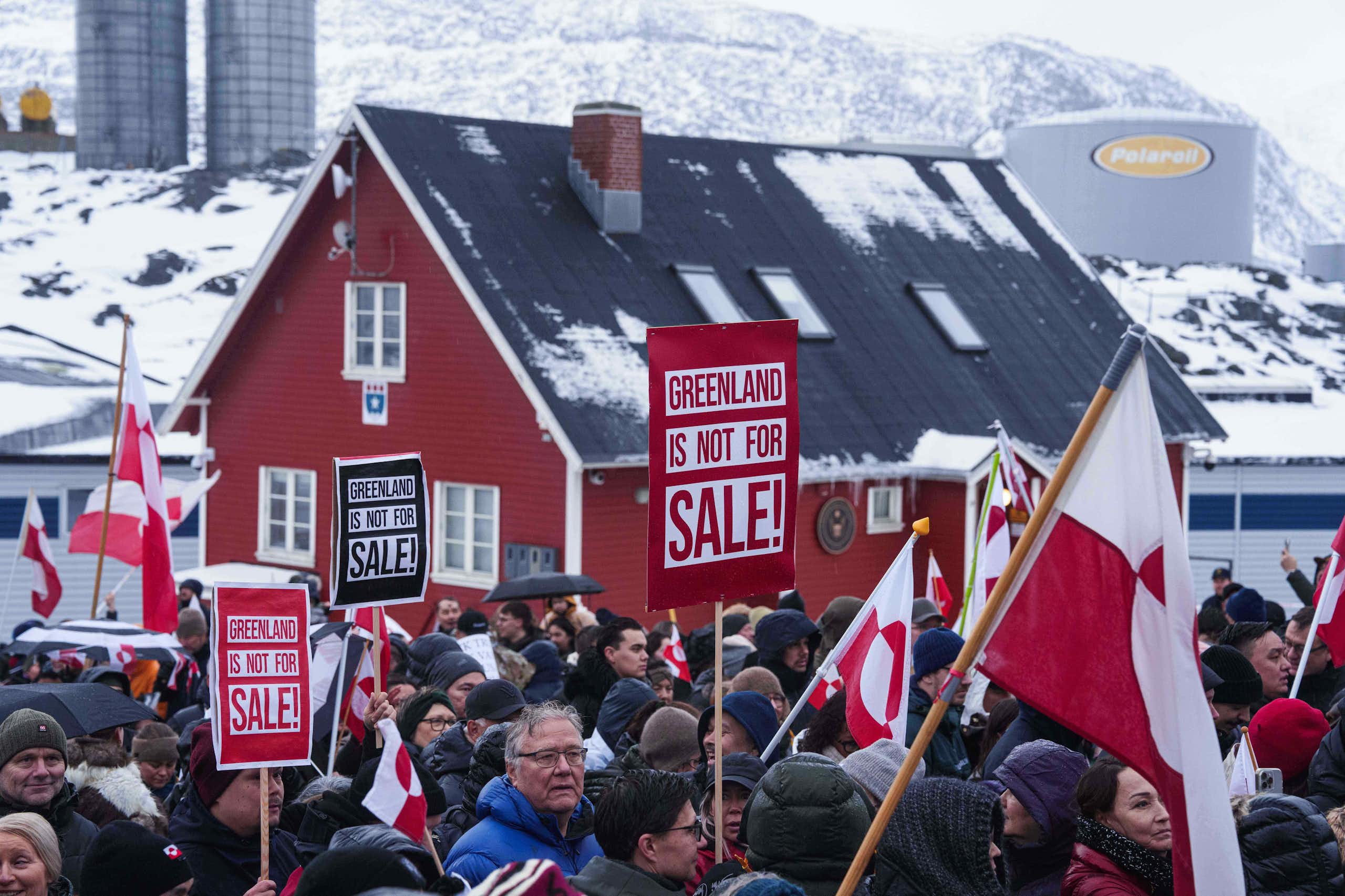 People in parkas protest in front of a red clapboard building holding signs that read Greenland is Not For Sale. Snowy hills are in the background.