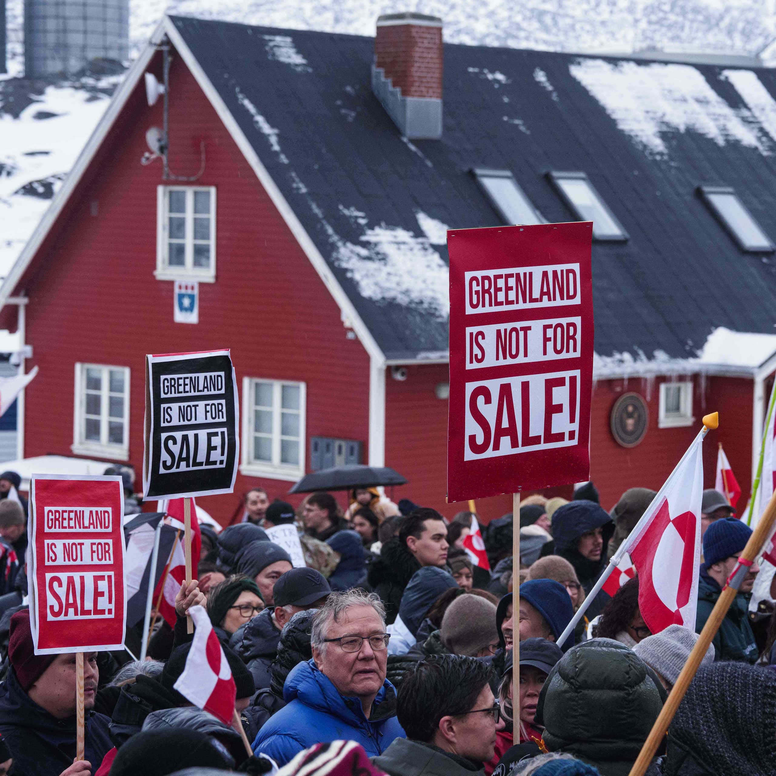 People in parkas protest in front of a red clapboard building holding signs that read Greenland is Not For Sale. Snowy hills are in the background.